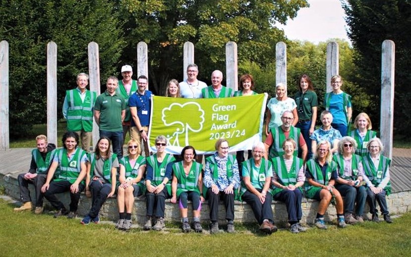 A group of volunteers from the Friends of Horsham Park, all wearing green shirts, stand holding a banner which reads "Green Flag Award 23/24" in front of a backdrop of trees.